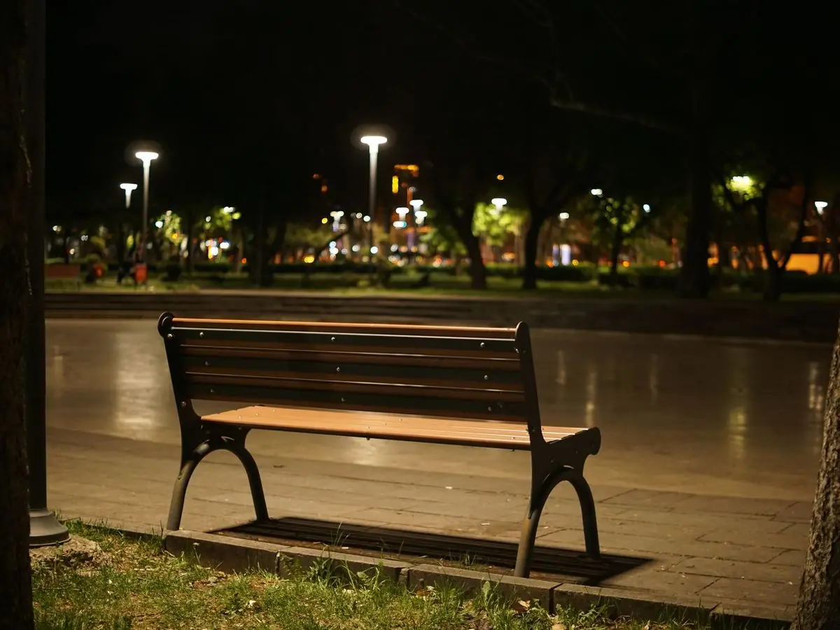An empty park bench at night, with city lights behind it.