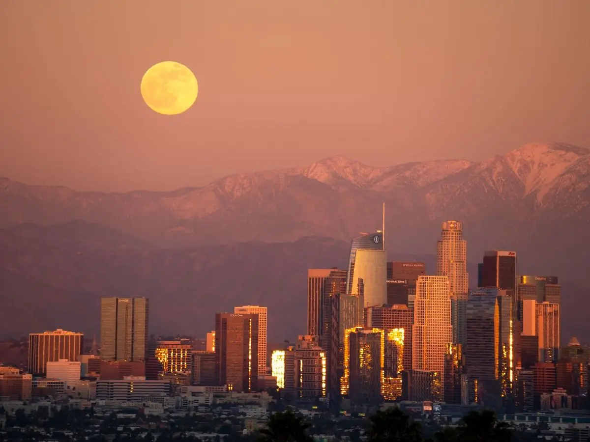 The downtown Los Angeles skyline at dusk, with a full moon over the mountains.